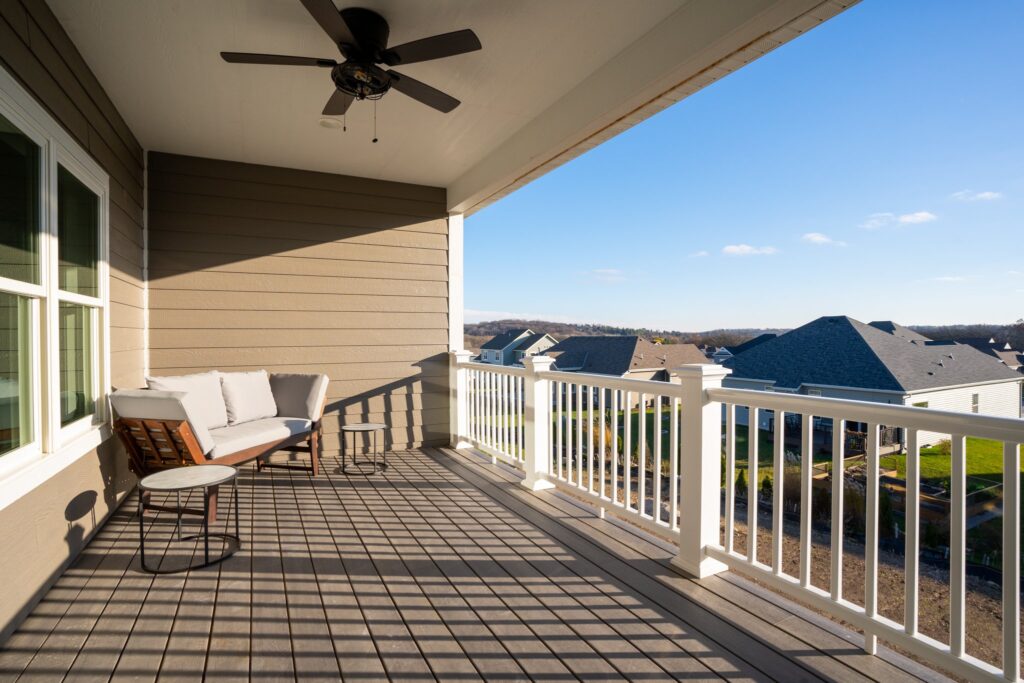 A spacious balcony with a ceiling fan, modern seating, and tables overlooks a serene suburban neighborhood on a clear, sunny day.