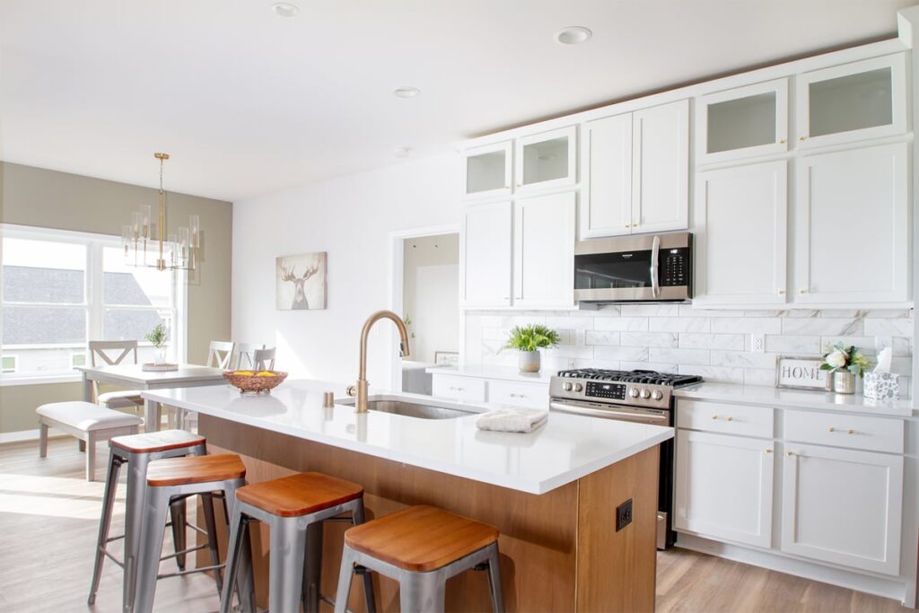 Bright modern kitchen with white cabinets, central island, barstools, and dining area. Decor includes wall art and plants. Sunlit space, elegant design.