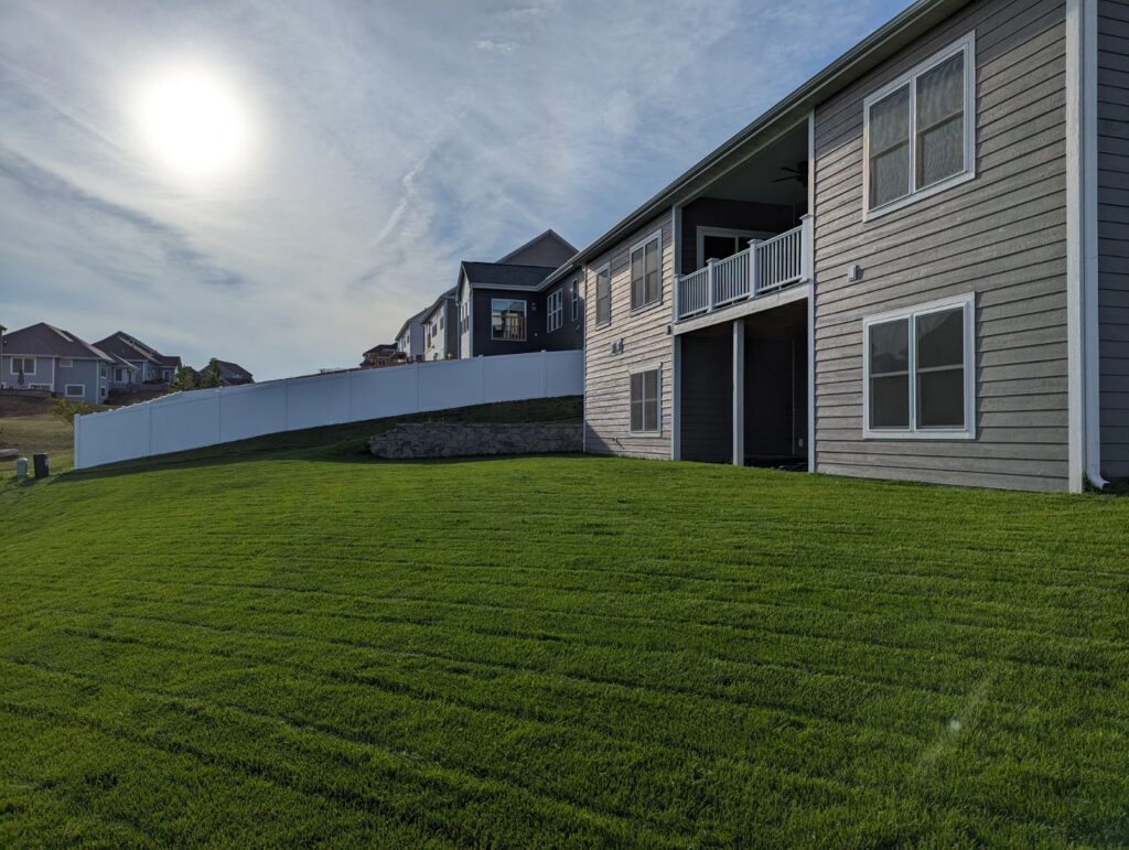 A suburban neighborhood with modern houses, green lawns, and a bright sky. The sun is prominently visible, casting long shadows.
