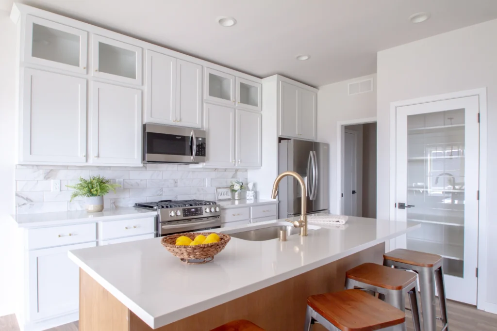 Modern kitchen with sleek white cabinets, marble backsplash, stainless steel appliances, and a central island featuring a gold faucet with stools and a fruit basket.