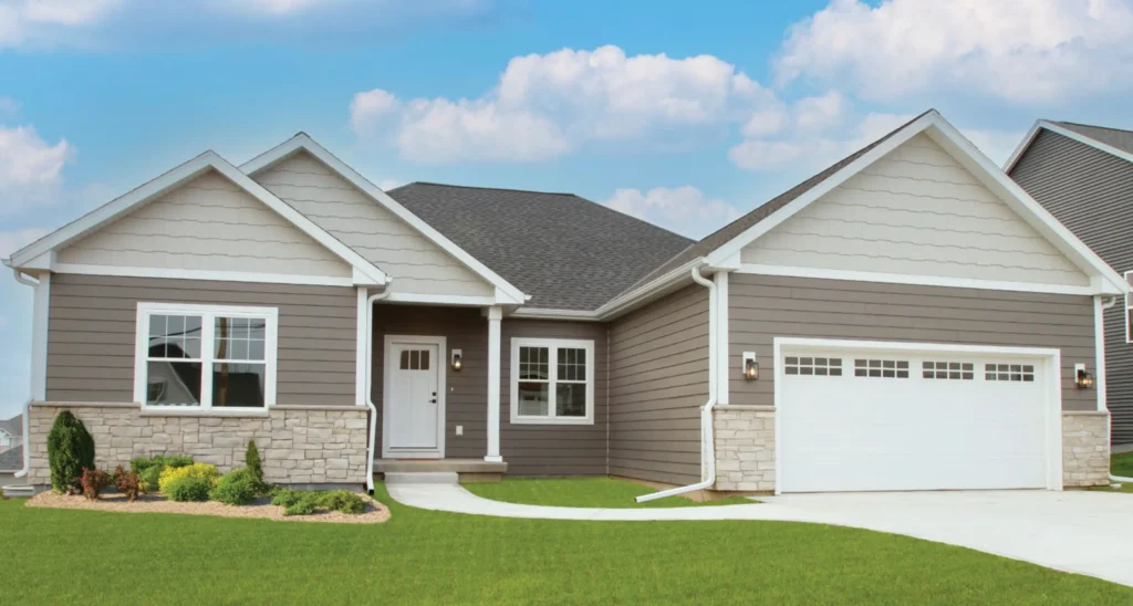 Single-story house with gray siding, a double garage, and landscaped front yard. Clear blue sky background. No recognizable landmarks or people visible.