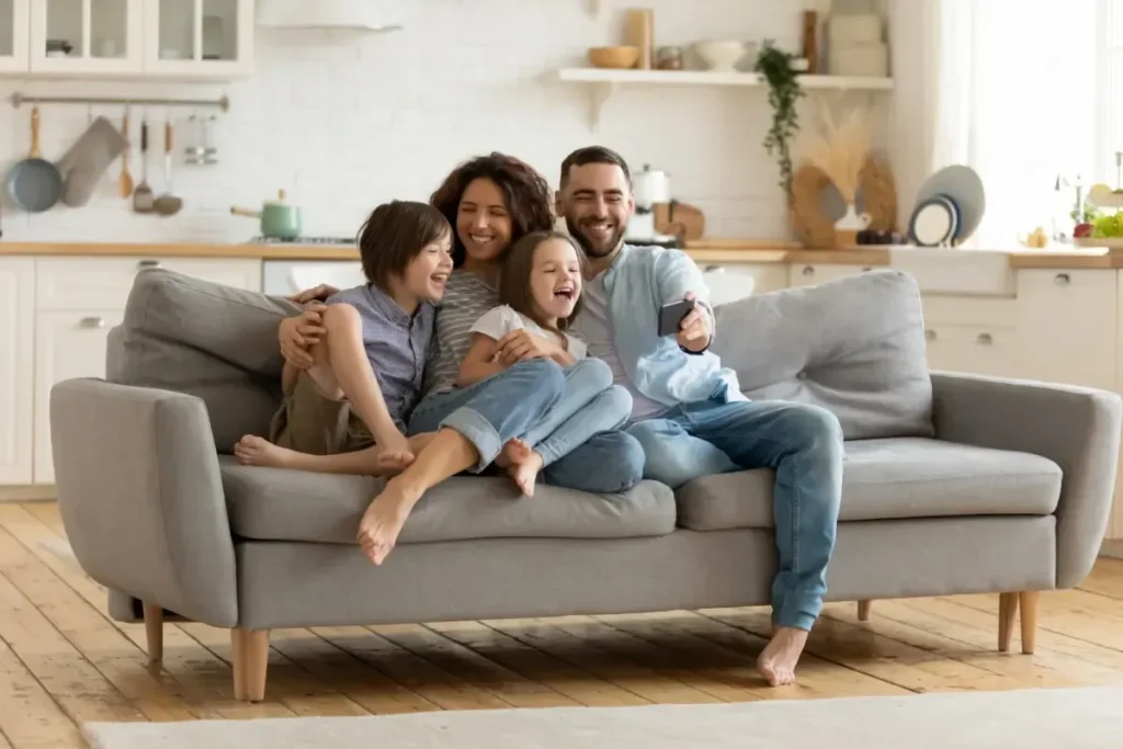 A joyful family sits on a gray sofa in a cozy, modern living room, smiling at a phone screen, surrounded by kitchen decor.
