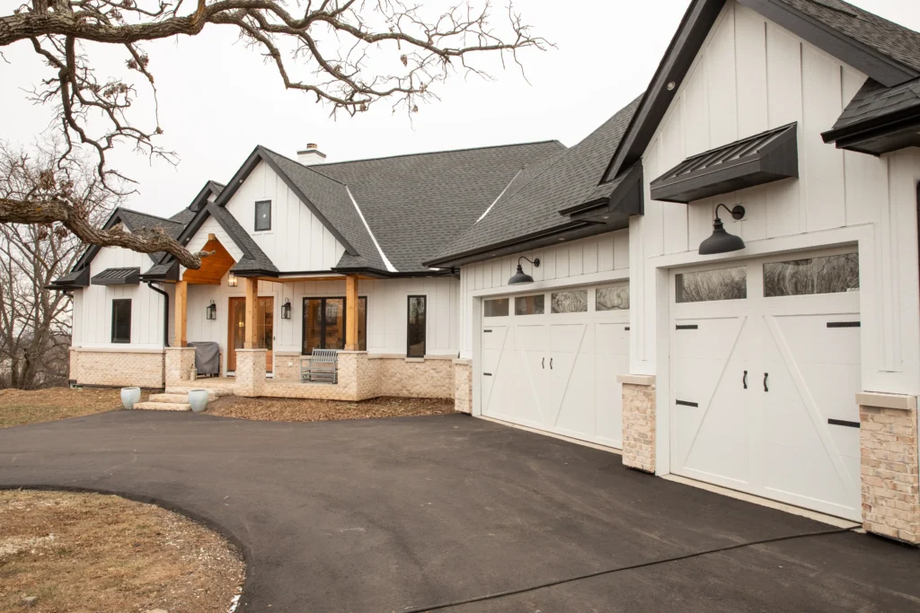 A modern suburban house with white siding, black roof, spacious garage, and a cozy front porch, surrounded by leafless trees in a quiet neighborhood.