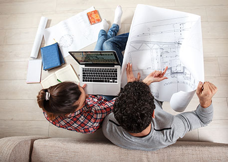 Two people reviewing architectural plans and a laptop on the floor, surrounded by books and papers in a workspace setting.